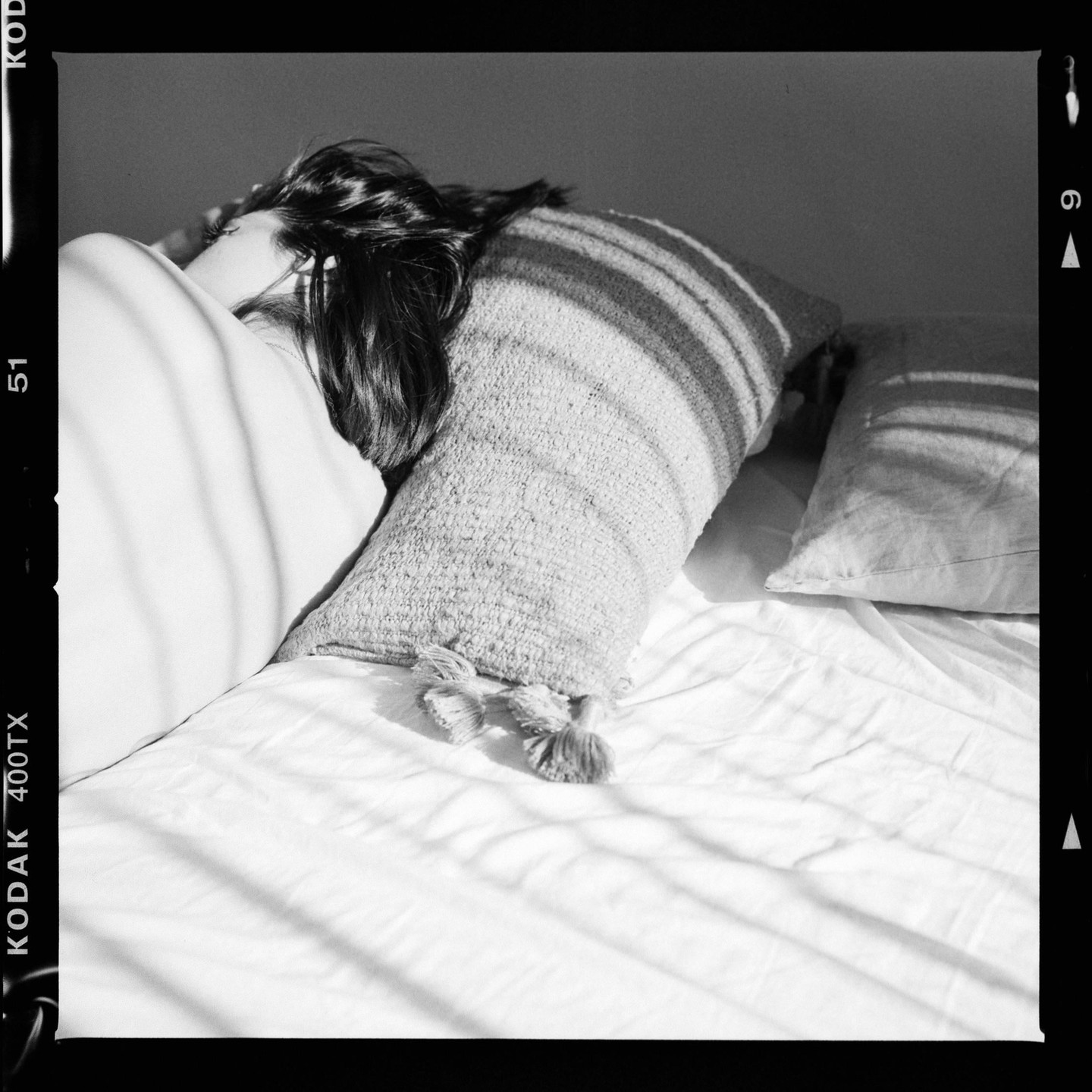 A woman lies on a bed with pillows, with shadows cast across the sheets.