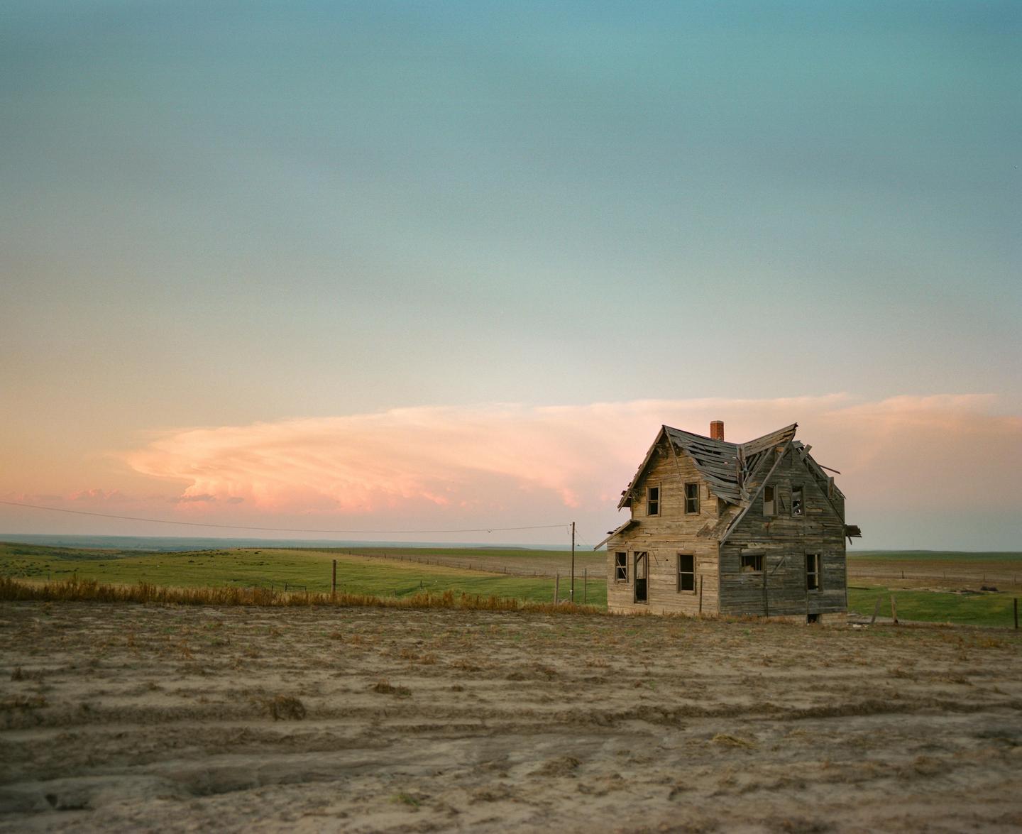 An abandoned wooden house with a damaged roof stands in a field under a sky with pink clouds.
