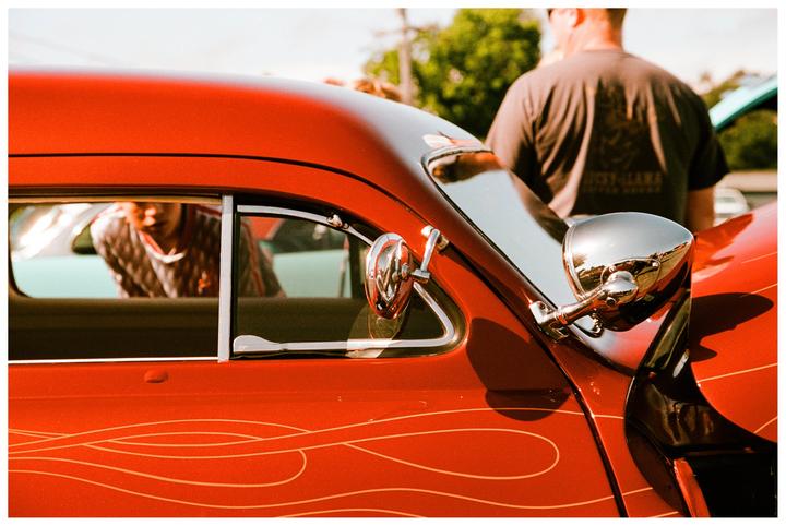A red vintage car with custom pinstriping is parked outdoors, with a man standing behind it and another man reflected in its window.