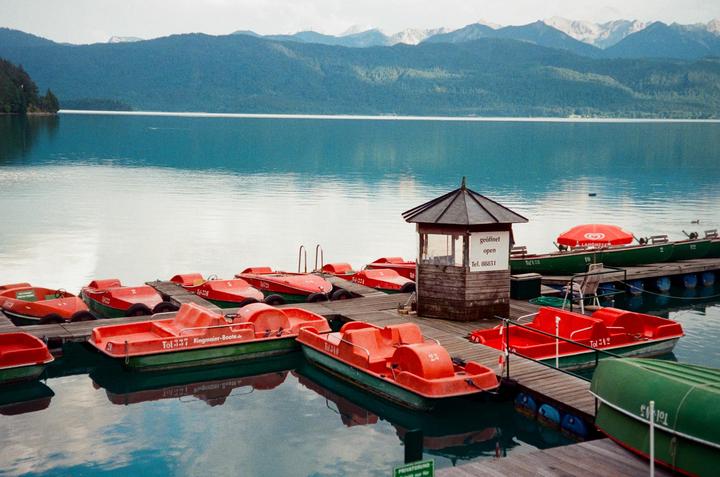 A wooden dock with red and green paddle boats and a small wooden hut sits on a calm lake surrounded by mountains.