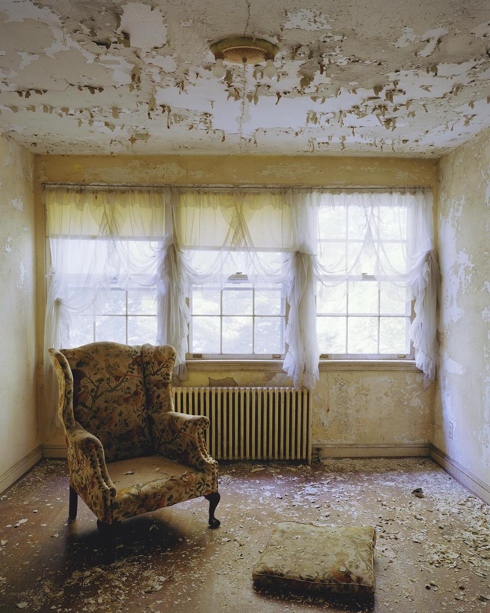 An abandoned room with peeling paint on the walls and ceiling contains a floral armchair, a footrest, and three windows with sheer white curtains.