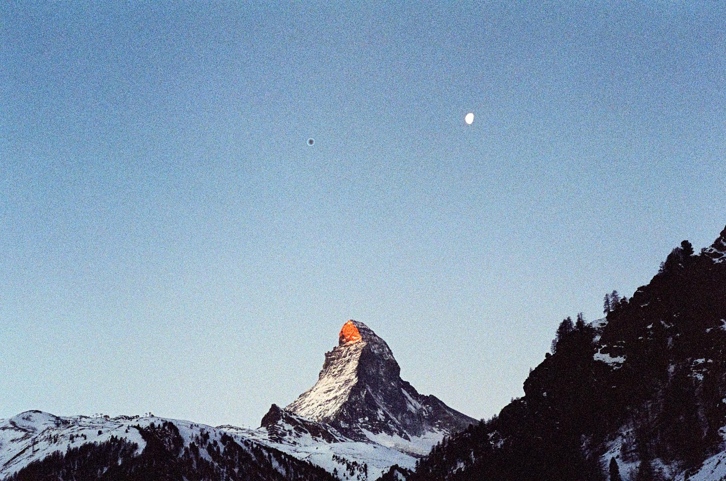 The Matterhorn mountain peak is illuminated by the setting sun, with a bright moon and a dark spot visible in the clear sky above.
