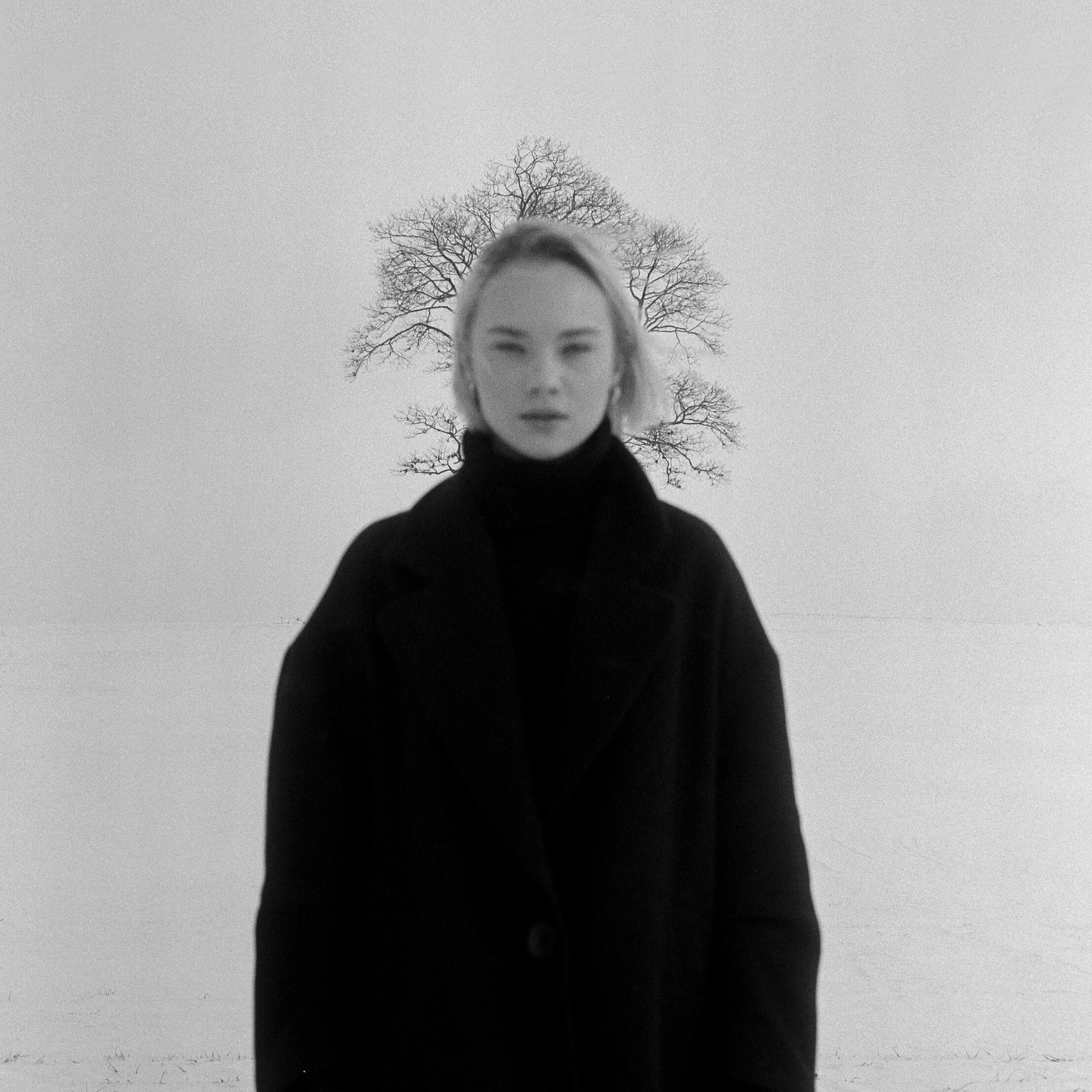 A woman in a dark coat stands in a snowy field with a bare tree behind her.