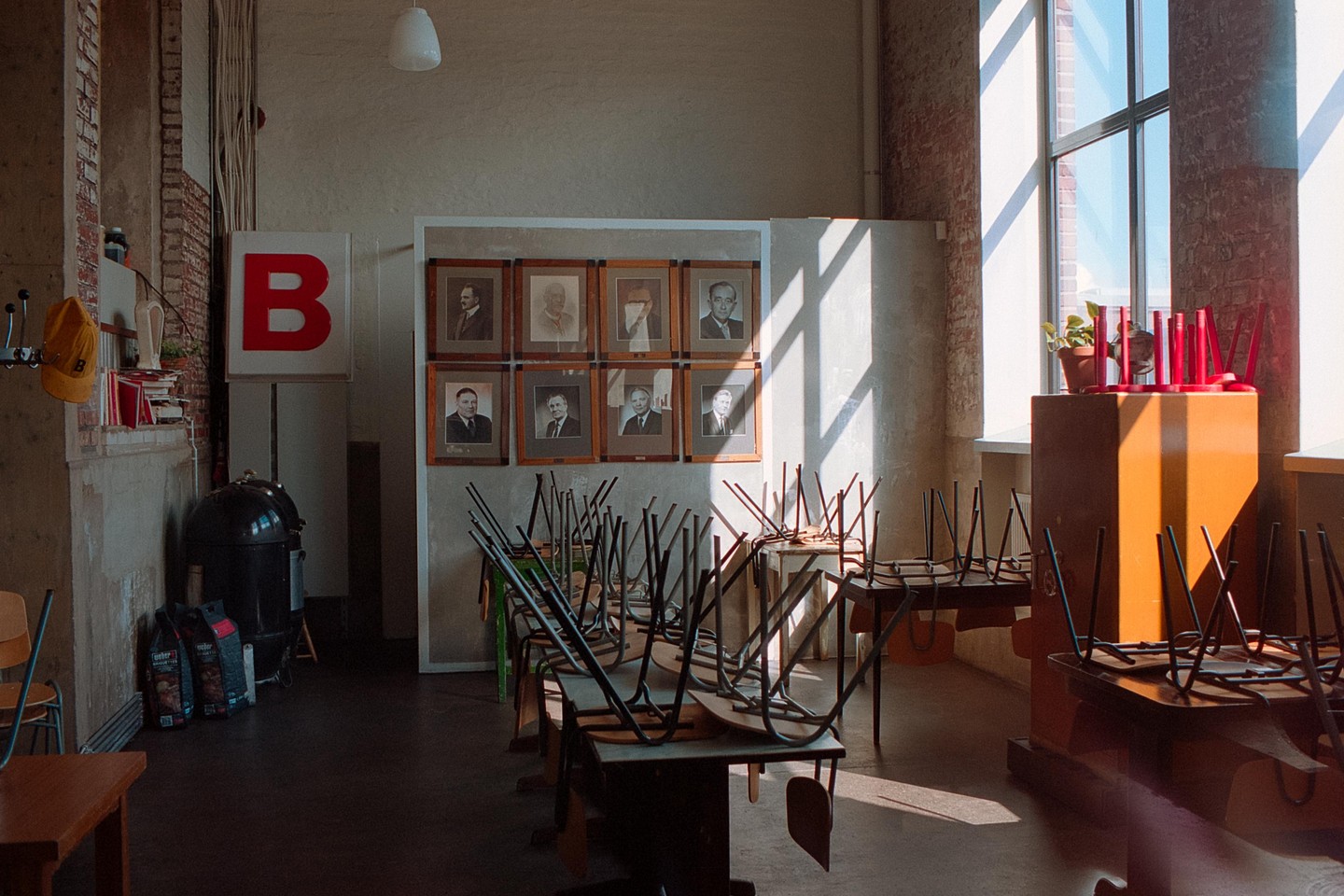 Sunlight streams through a large window onto tables with chairs stacked upside down in a room with framed portraits on the wall.
