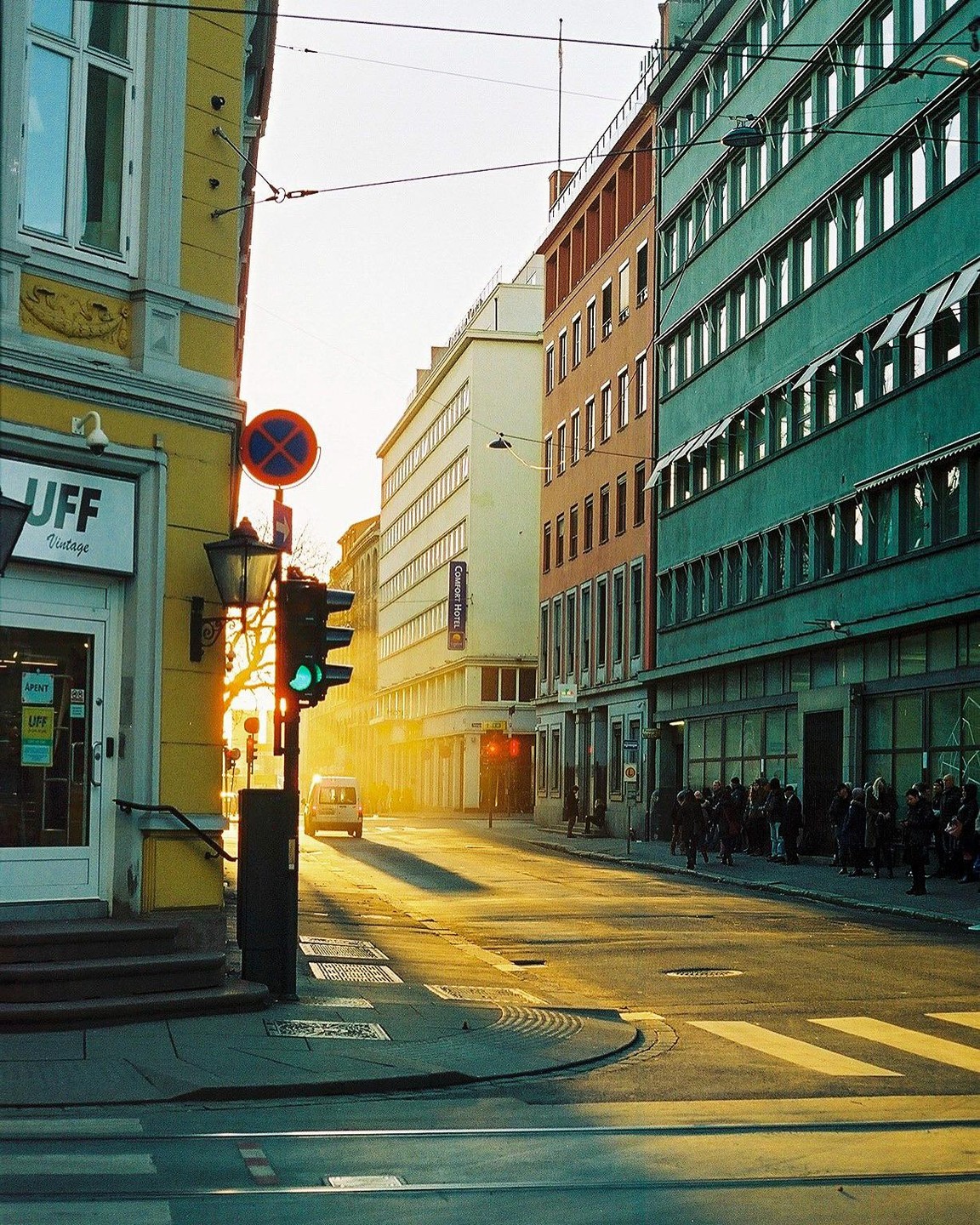 A street in Oslo is illuminated by the golden light of a sunset, with buildings lining both sides and people waiting on the sidewalk.