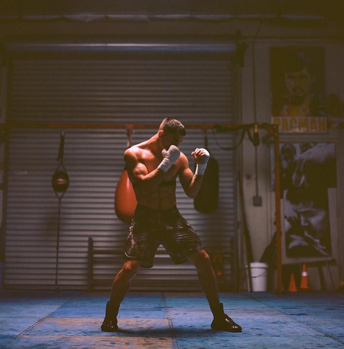 A man in boxing stance is training in a gym with punching bags and a poster in the background.