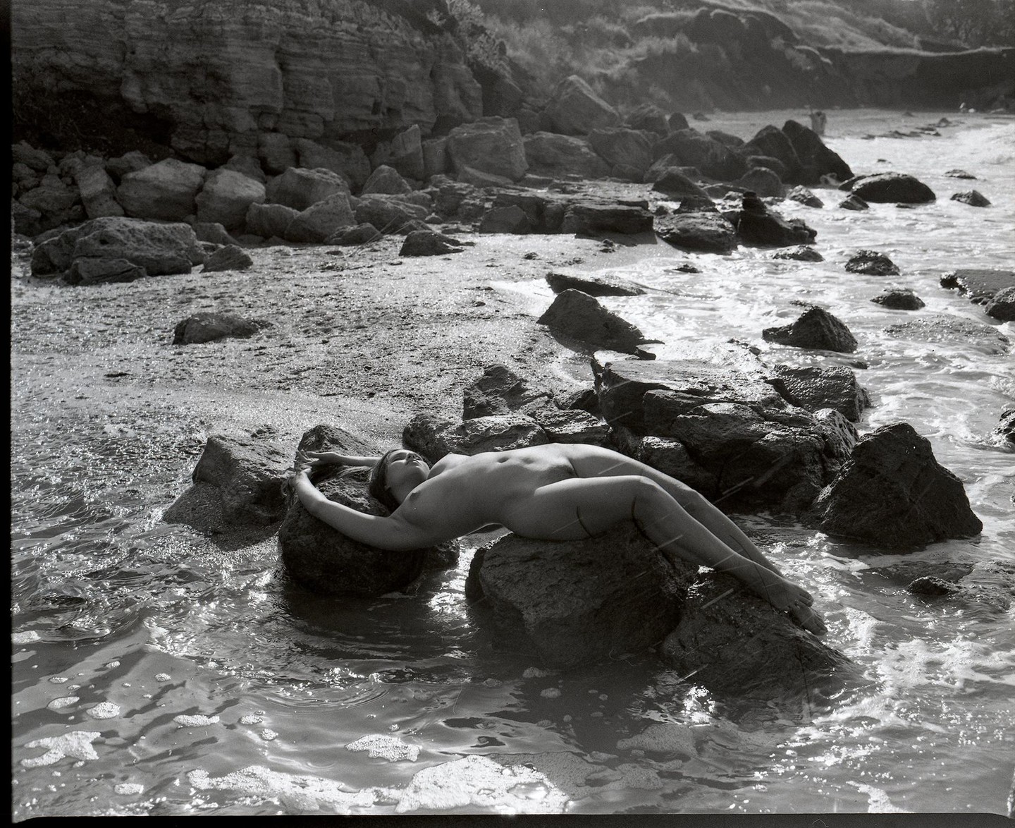 A nude woman lies on a rock in the water at a rocky beach.