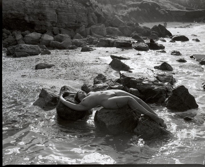 A nude woman lies on a rock in the water at a rocky beach.