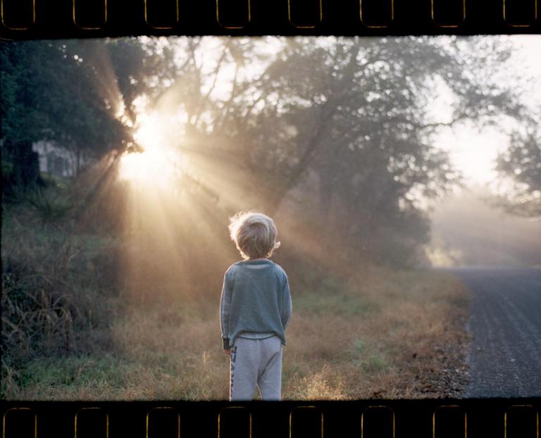 A child stands on the edge of a road, looking towards a bright sunburst through trees.