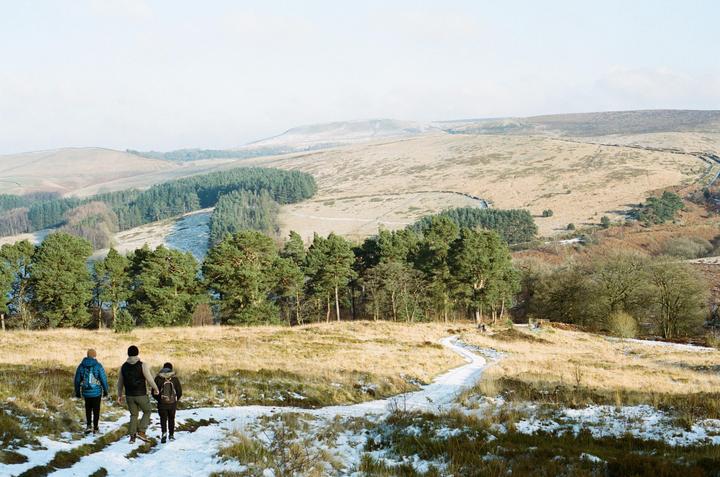 Three people walk on a snowy path through a winter landscape with hills and trees.