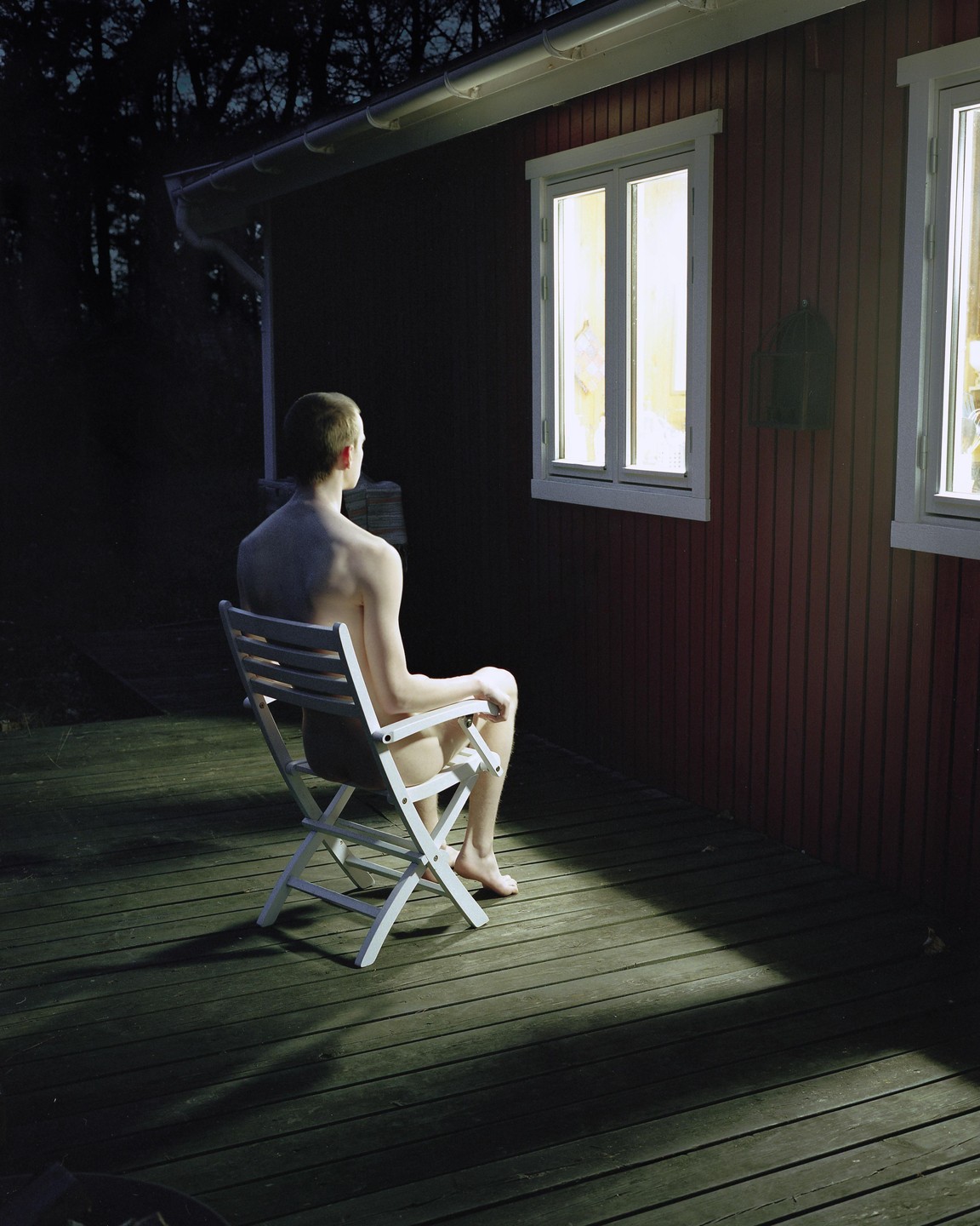 A man sits shirtless on a chair on a wooden deck at night, looking towards a brightly lit window of a red house.