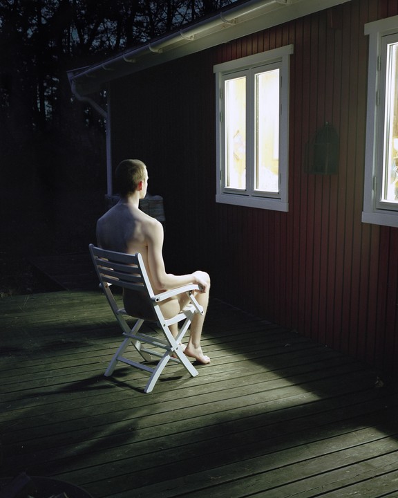 A man sits shirtless on a chair on a wooden deck at night, looking towards a brightly lit window of a red house.