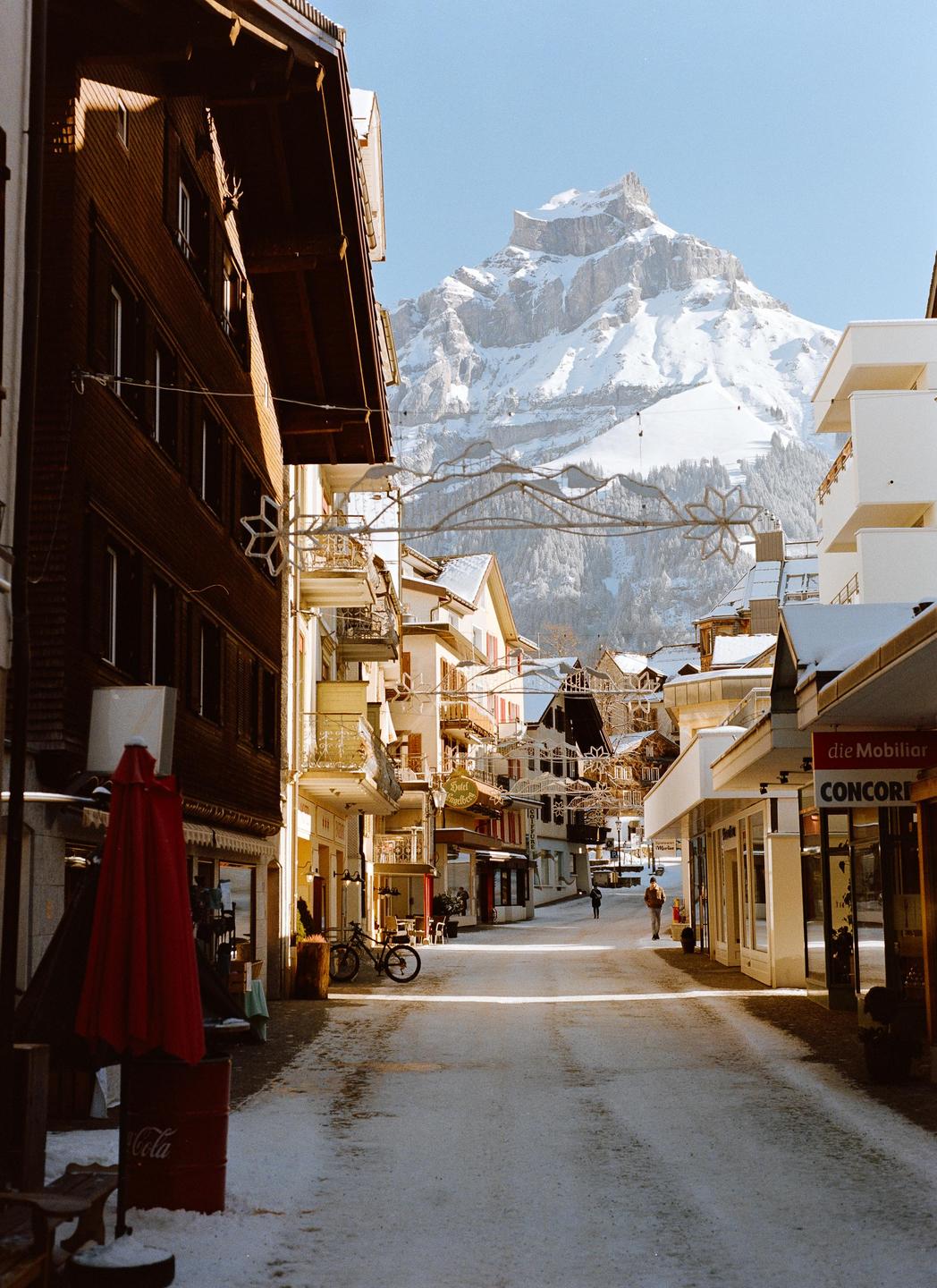 A snow-covered street in a mountain town is lined with buildings and leads to a large snow-capped mountain.