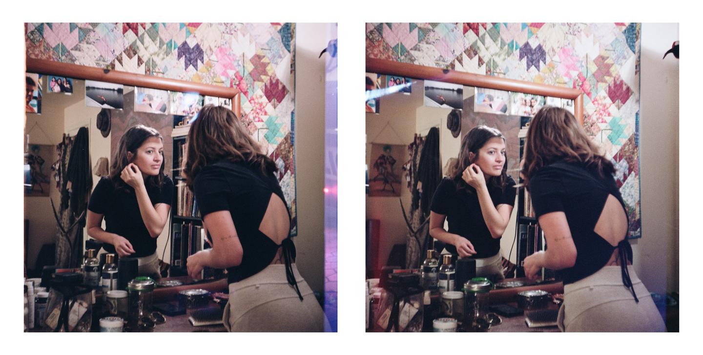 A woman looks at her reflection in a mirror while adjusting an earring, with a colorful quilt hanging on the wall behind her.