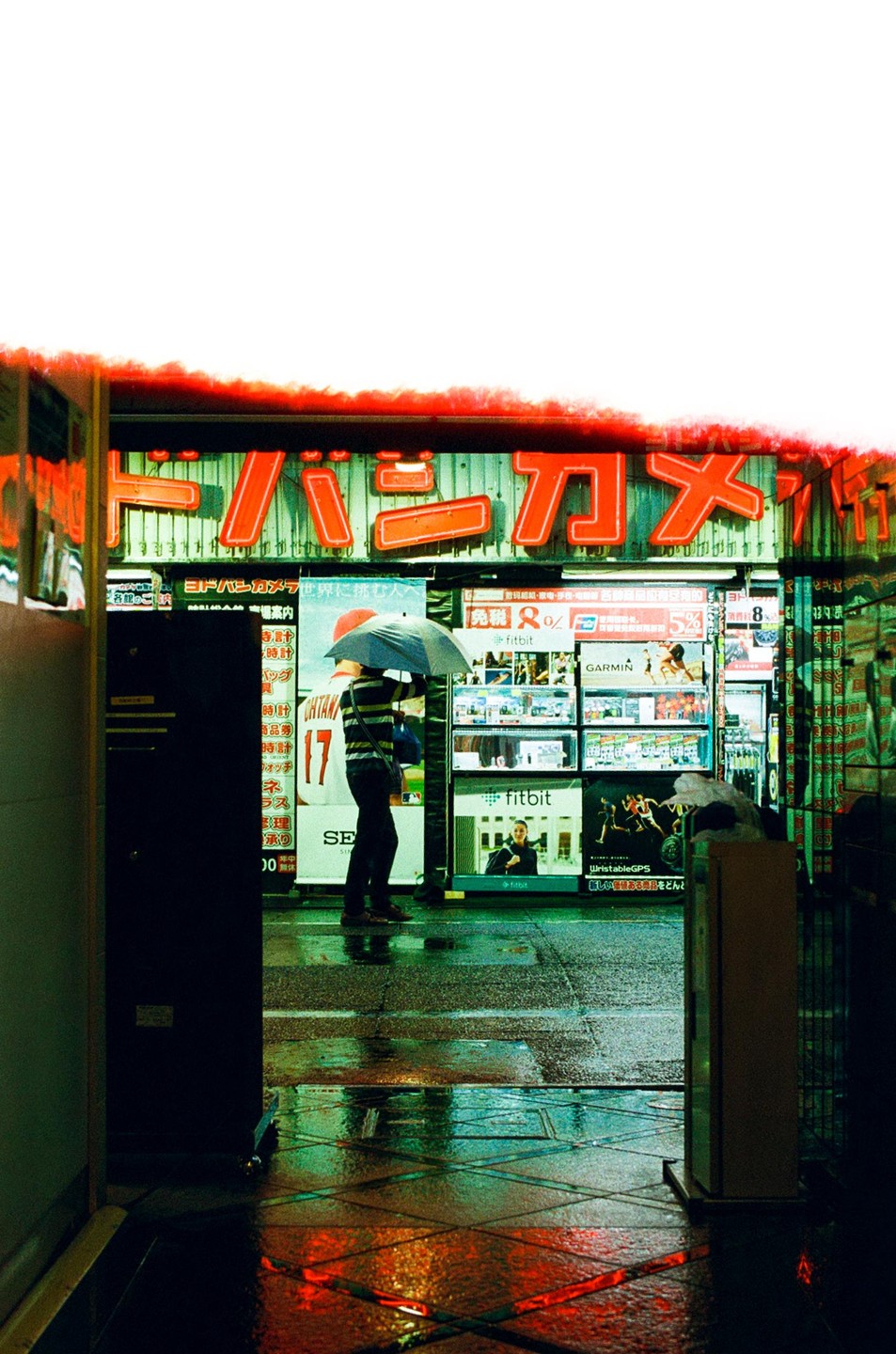 A person holding an umbrella stands on a wet street in front of a store with bright neon signs.