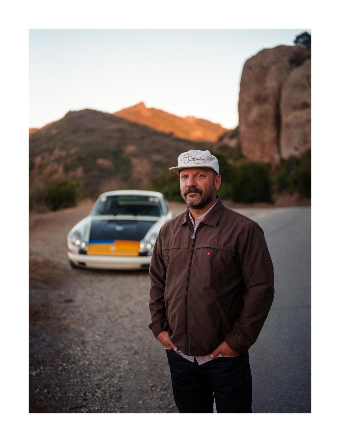 A man stands on a road with a car behind him and mountains in the background.
