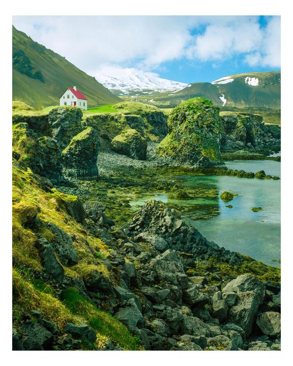 A white house with a red roof sits on a green hill overlooking a rocky coastline with clear water and snow-capped mountains in the background.