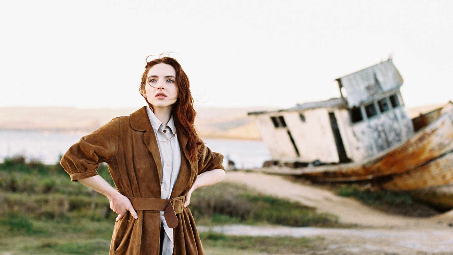 A woman stands in front of a shipwreck on a beach.