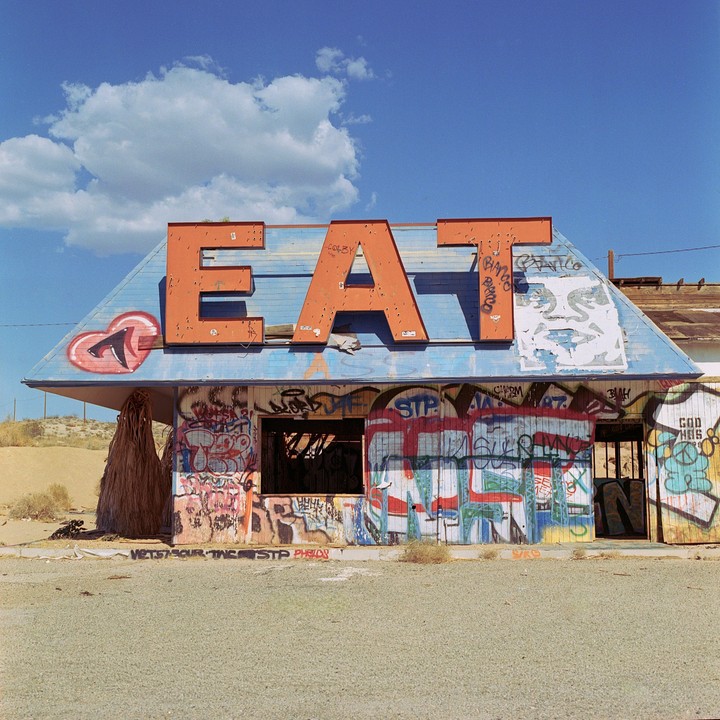 An abandoned building with a blue roof and the word "EAT" in large orange letters on top is covered in graffiti under a bright blue sky with a single cloud.