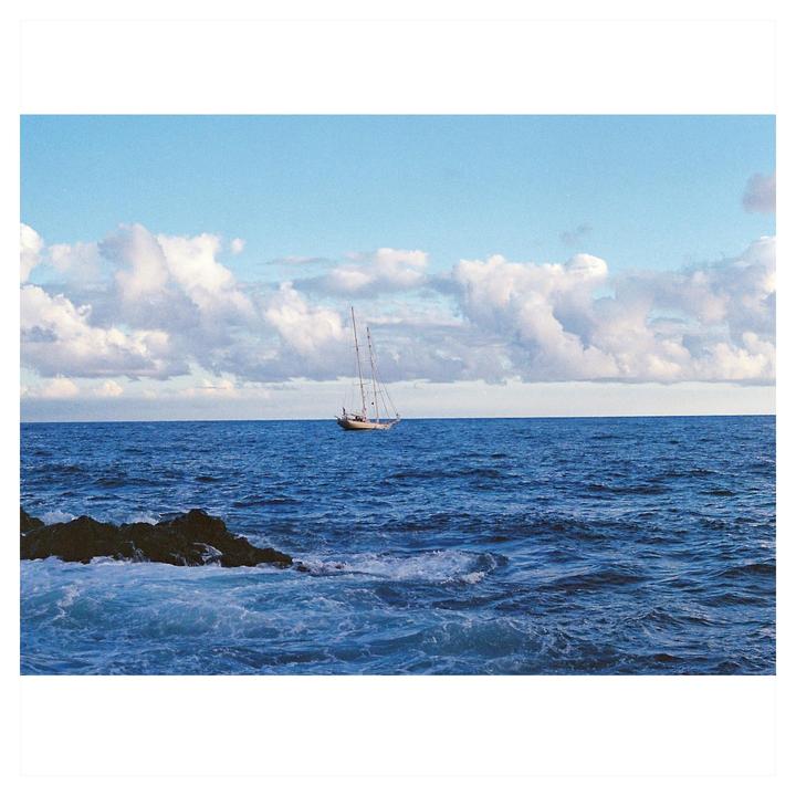A sailboat floats on the ocean with a rocky shore in the foreground under a cloudy sky.