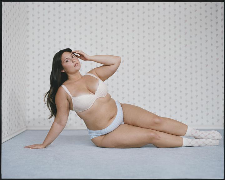 A woman in underwear and socks is posing on a carpeted floor against a patterned wall.