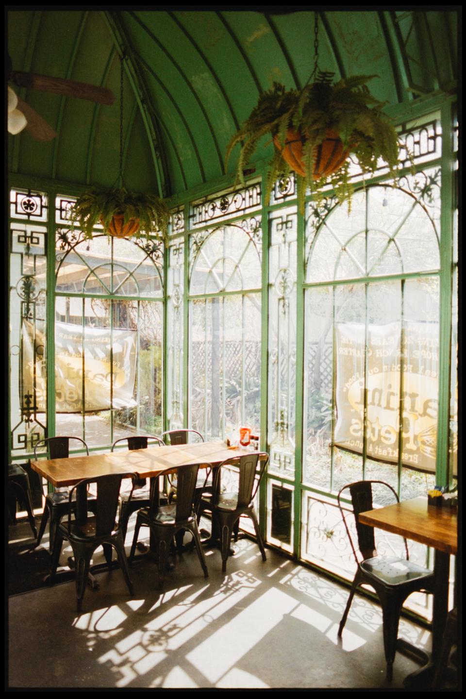A sunroom with green window frames, tables, and chairs is filled with bright sunlight and shadows.