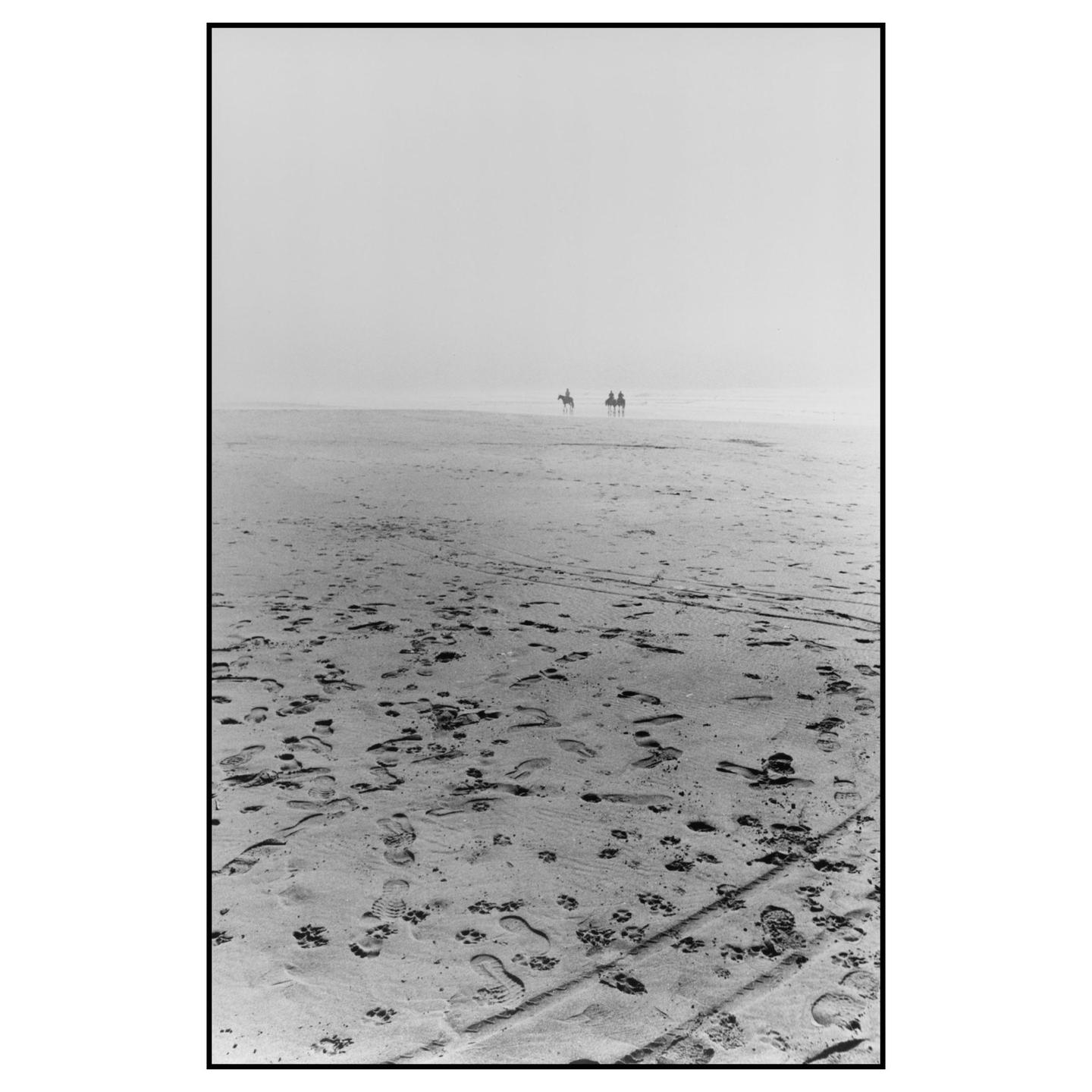 Two people on horseback are visible in the distance on a hazy beach covered in footprints.