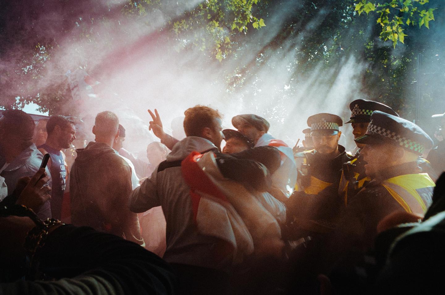 A crowd of people, including police officers, are gathered in a hazy, sunlit outdoor setting with trees overhead.