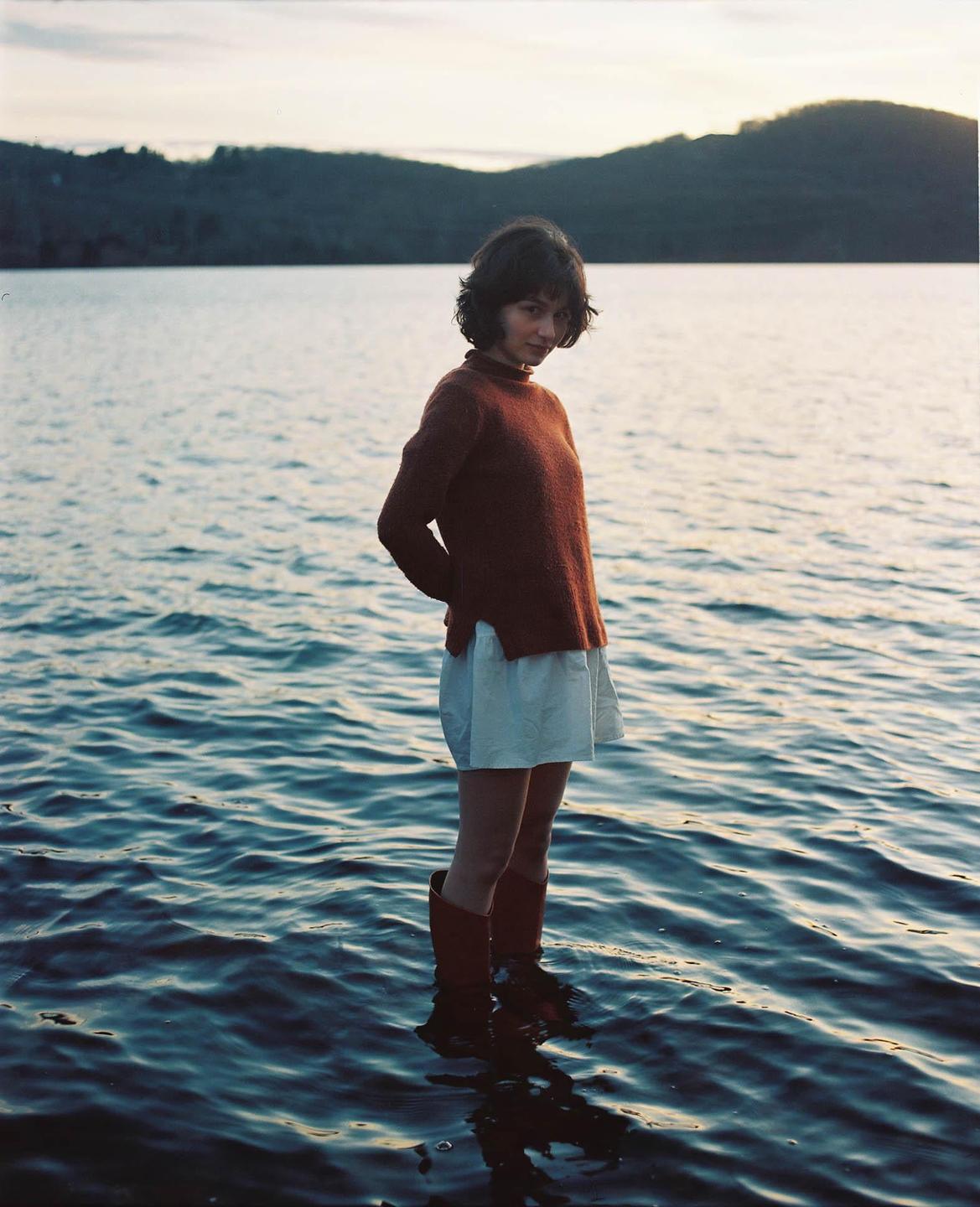 A woman stands in a body of water with mountains in the background.