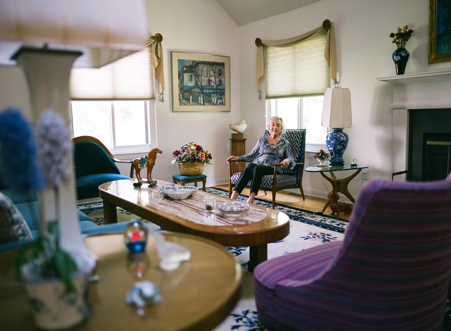 A woman sits in an armchair in a living room with a coffee table and various decorations.