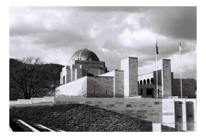 A large domed building with flags flying in front of it stands on a hill with trees and a cloudy sky in the background.