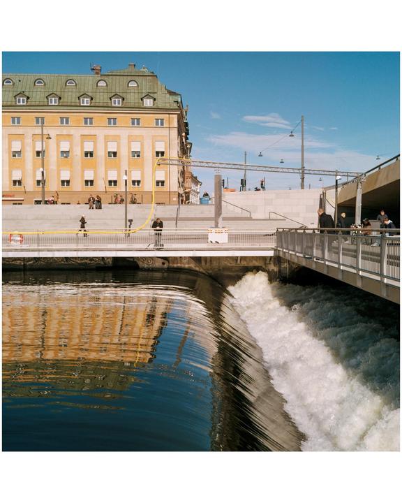 A building with a green roof is reflected in the water next to a waterfall and a bridge with people.