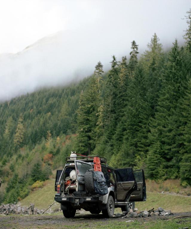 A vehicle with its doors open and camping gear loaded on the roof and in the back is parked on a dirt road in a forest with a hazy mountain in the background.