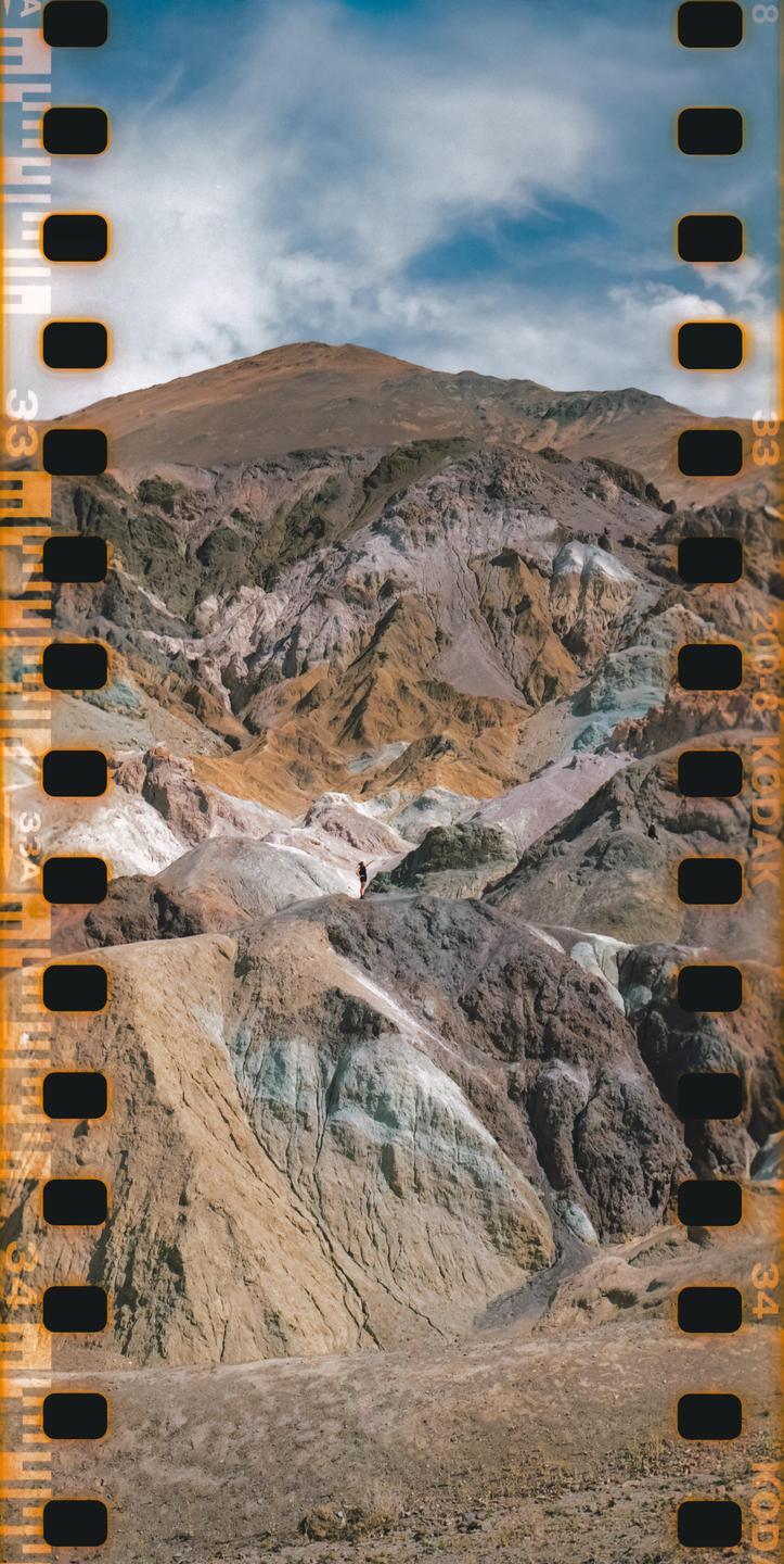 A person stands on a ridge overlooking a vast, colorful mountainous landscape under a partly cloudy sky.