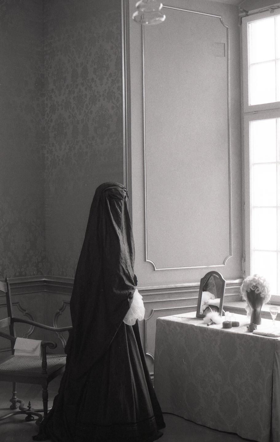 A person in a dark, flowing garment stands next to a dressing table with a mirror and a wig.