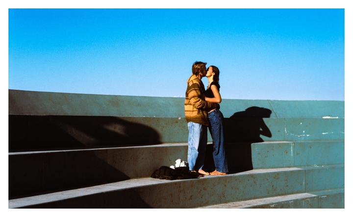 A young couple is kissing on concrete steps under a clear blue sky.