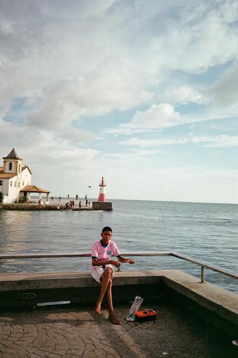 A boy sits barefoot on a concrete ledge by the water, holding a fishing line, with a white building and a red and white lighthouse in the background.
