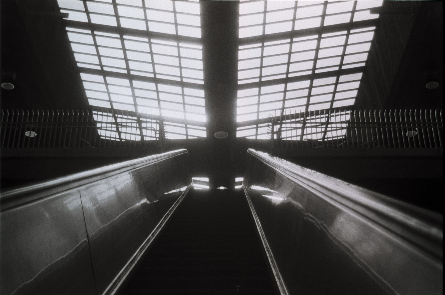 An upward-looking view of an escalator shows a bright, gridded ceiling and railings on either side.