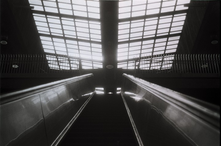 An upward-looking view of an escalator shows a bright, gridded ceiling and railings on either side.