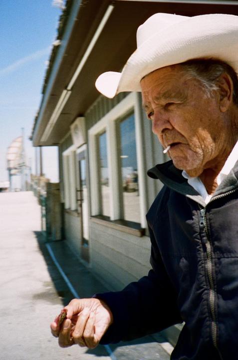 A man wearing a cowboy hat and holding a cigarette in his mouth stands next to a building on a sunny day.