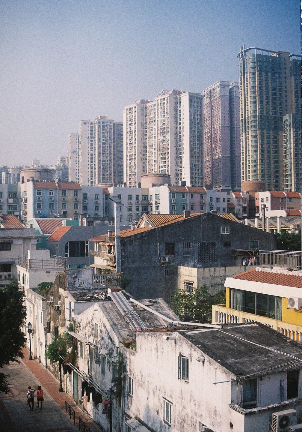 A street with old buildings and two people walking is in the foreground, with modern high-rise buildings in the background.