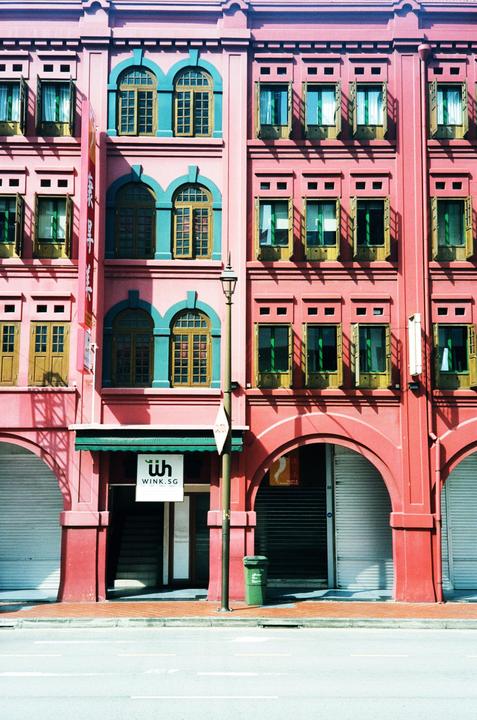 A pink building with teal window frames and arches stands next to a street with a green trash can and a lamppost.