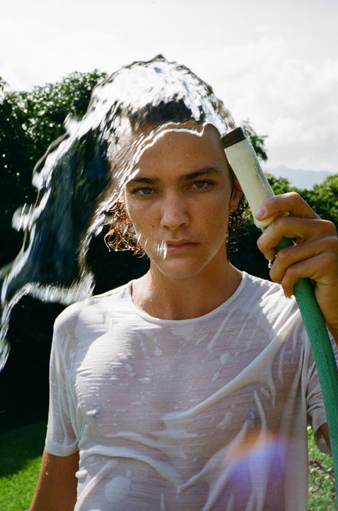 A man in a wet white shirt holds a garden hose as water pours over his head.