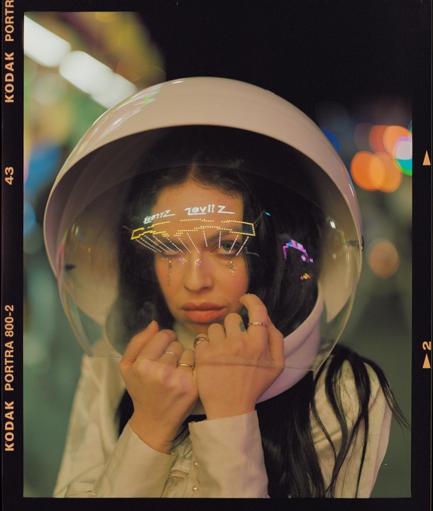 A woman wearing a white space helmet with reflections of colorful lights on the visor looks directly at the viewer.