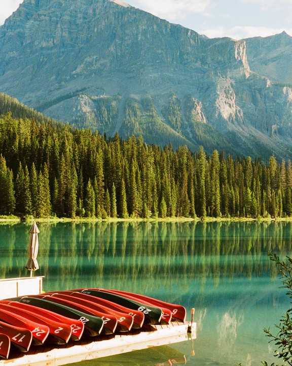 Red and green canoes are lined up on a dock in front of a lake with a forest and mountain in the background.
