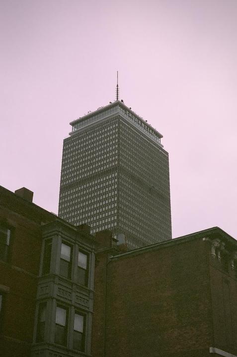 A tall skyscraper with a spire on top rises above brick buildings under a pink sky.
