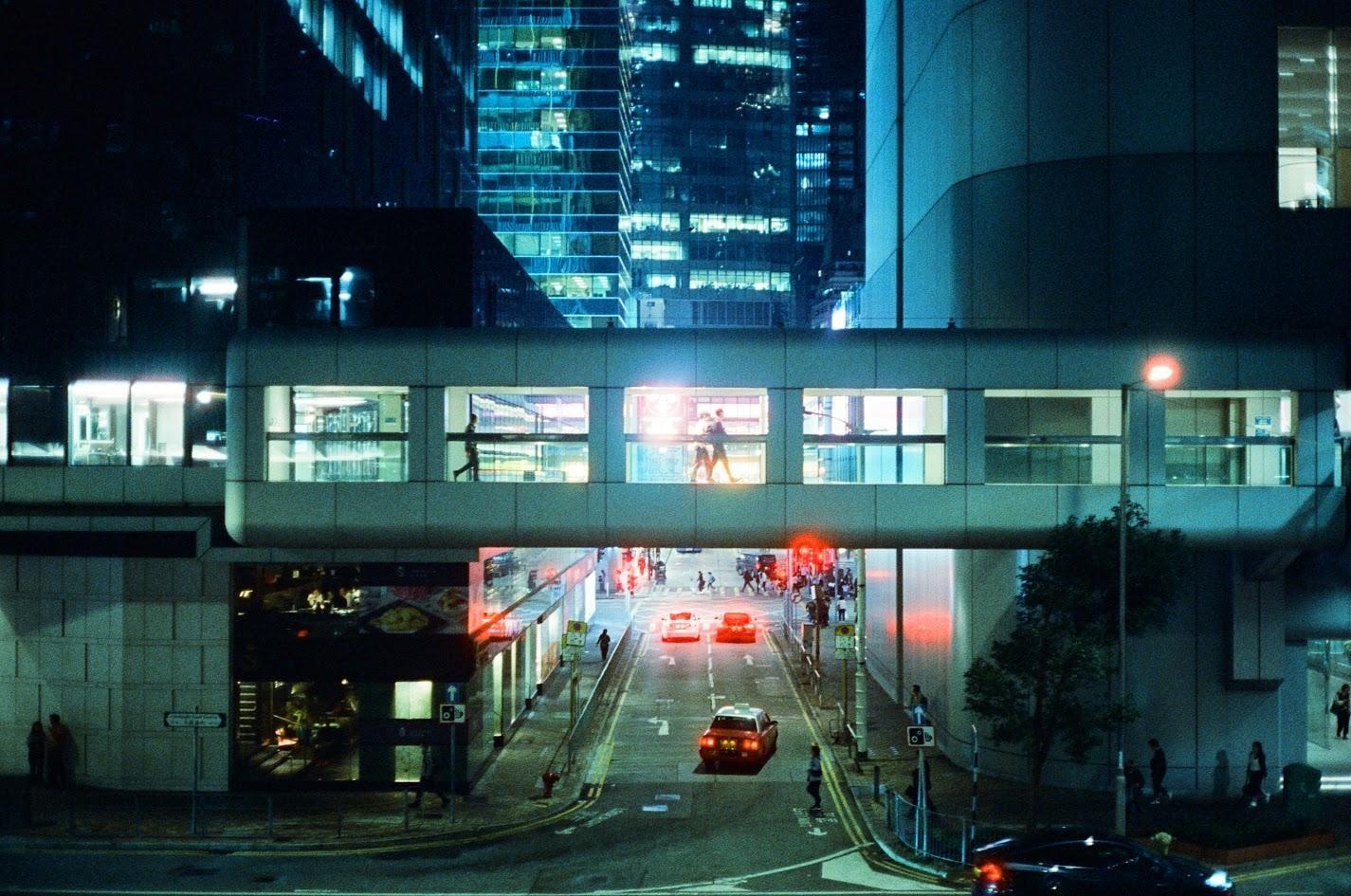 A street at night is viewed from above, with cars and people moving below a pedestrian bridge connecting two buildings.