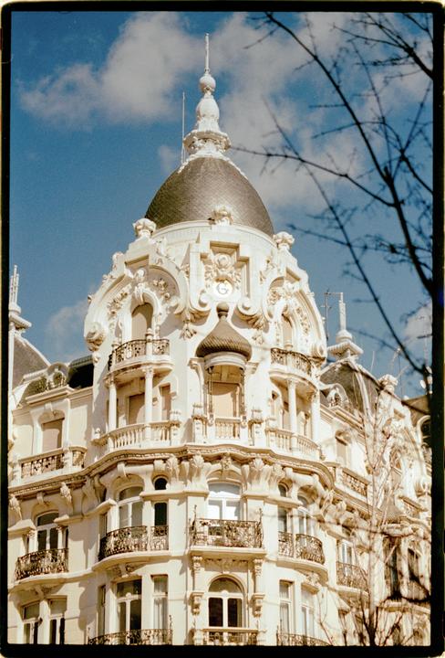 A white ornate building with a dark dome and balconies is visible against a blue sky with clouds.