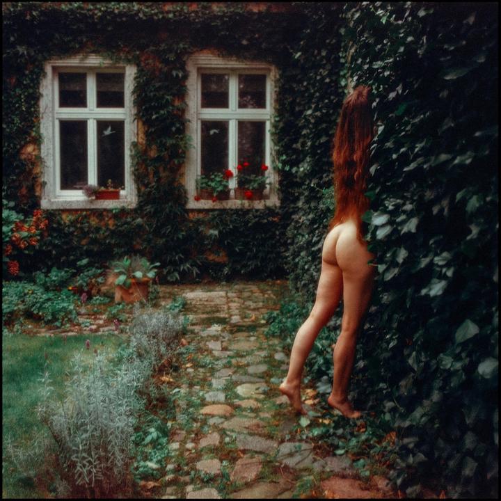 A nude woman with long red hair stands next to an ivy-covered wall with two windows, with a stone path and garden in the foreground.
