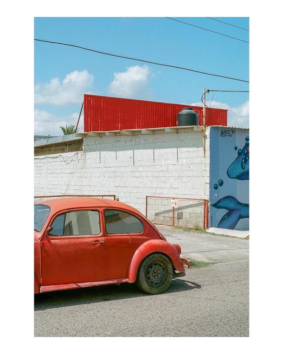 A red car is parked on a street next to a white wall with a blue mural and a red building under a clear sky.